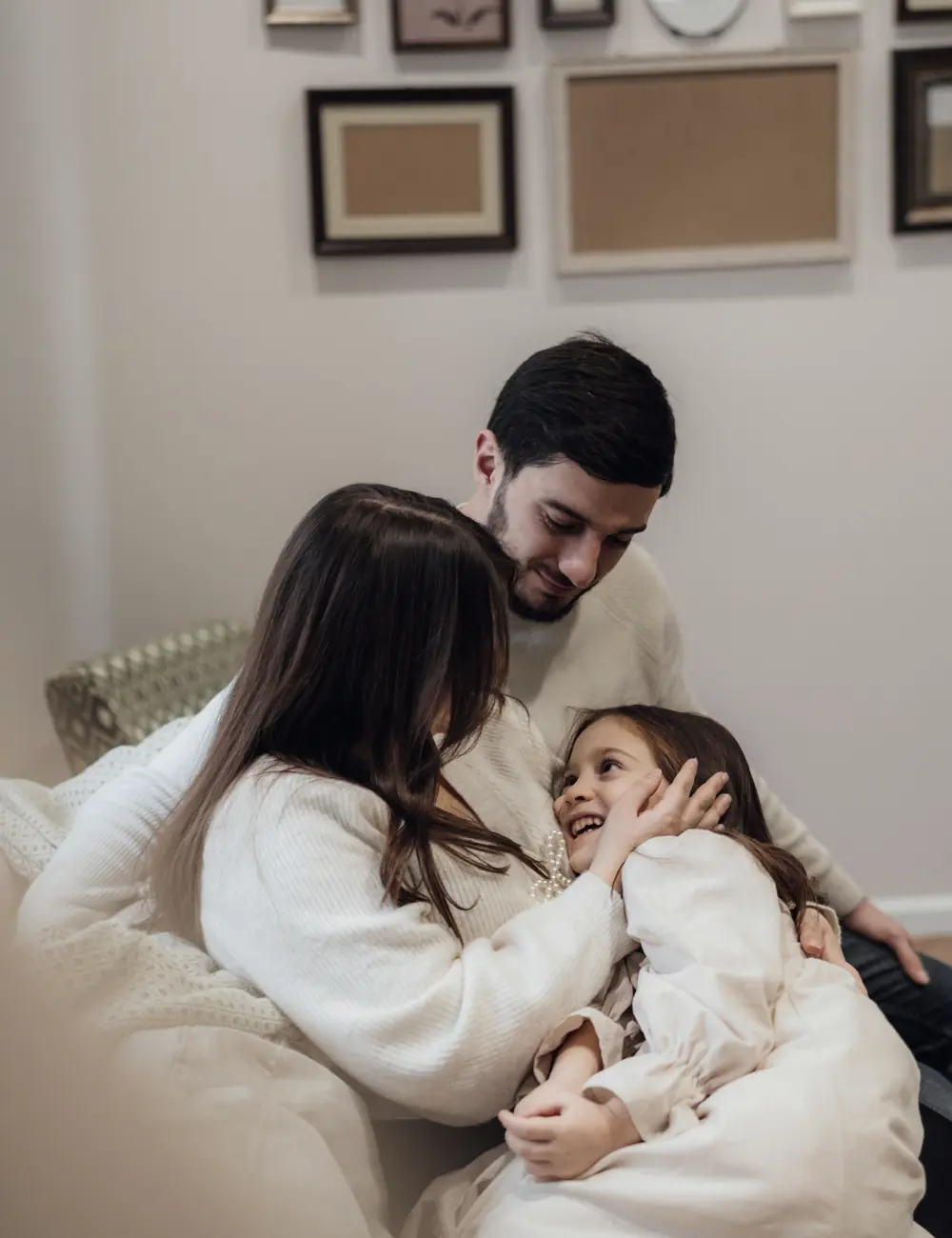 Pai, mãe e filha pequena abraçados e sorrindo em um sofá claro